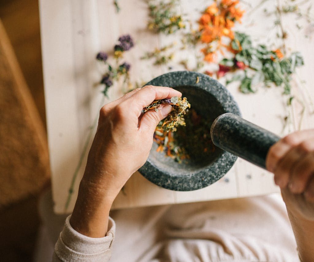 Top view of hands grinding dried herbs using a mortar and pestle on a wooden table.